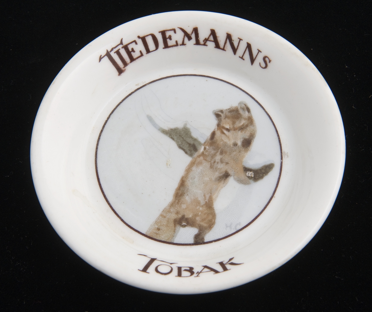 Liten, rund skål i porselen med reklame for Tiedemanns Tobak. Påtrykket motiv av en fjellrev etter original av Halfdan Gran.
