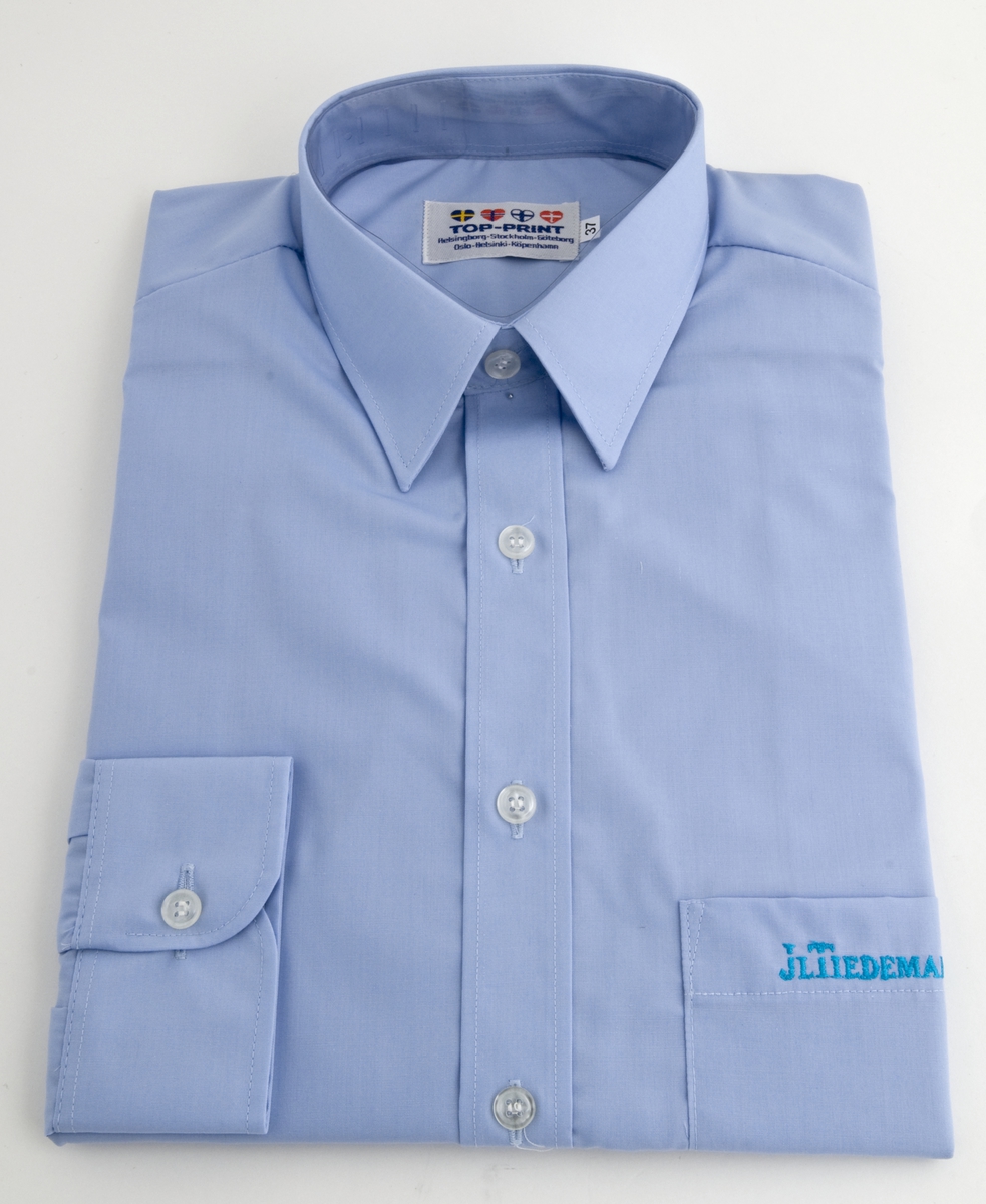Skjorte, lys blå med JL TIEDEMANNS i blått brodert med maskinsøm på venstre brystlomme.