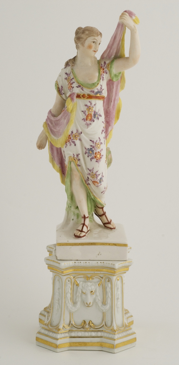 Figur; porselen, kvinne i antikiserende gevant på firesidet fotplate festet til postament.
Merket korslagte sverd på sokkelen.