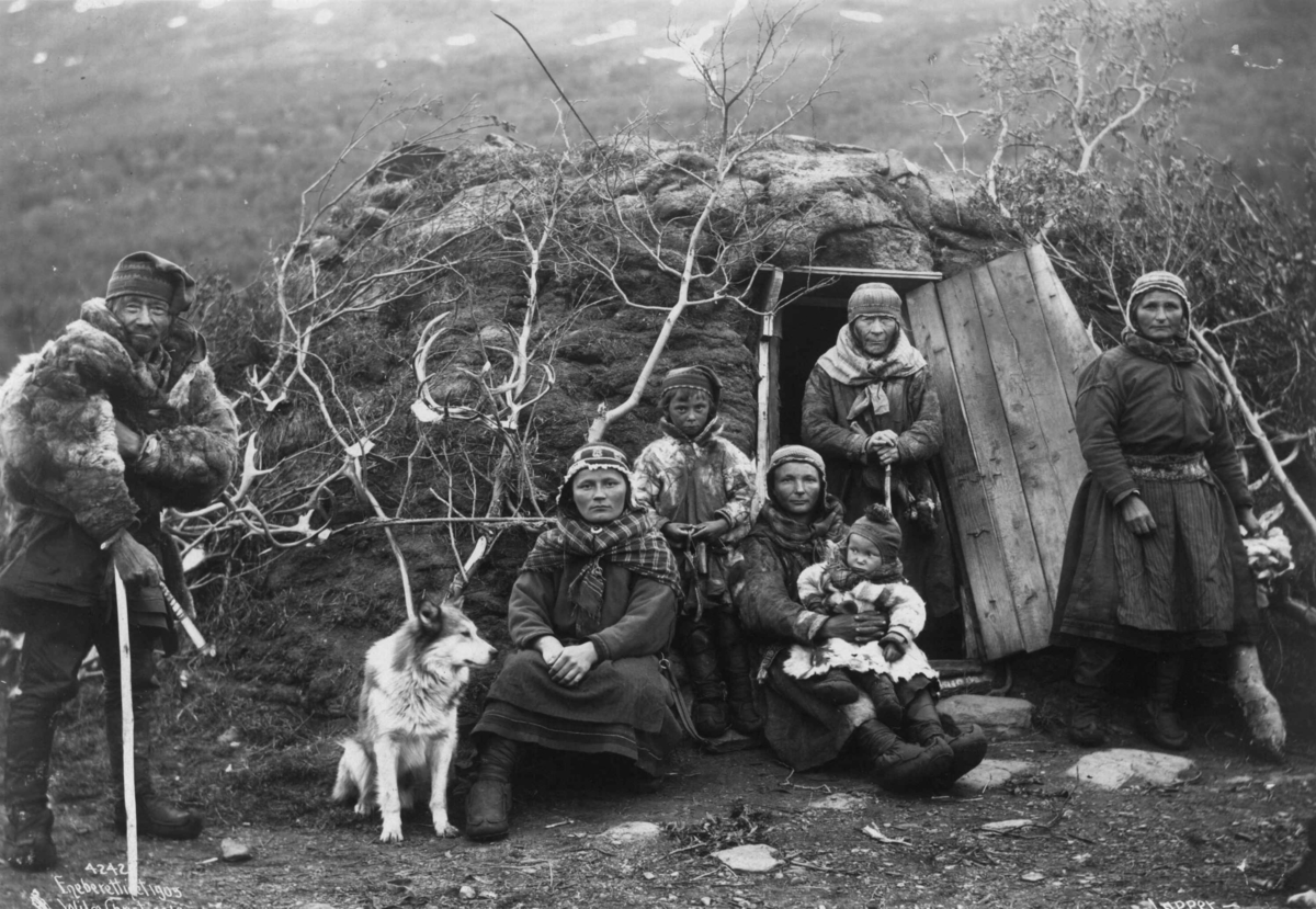 Svenske samer og en hund utenfor døråpningen til en gamme, fem voksne og to barn. Nord-Norge 1906.