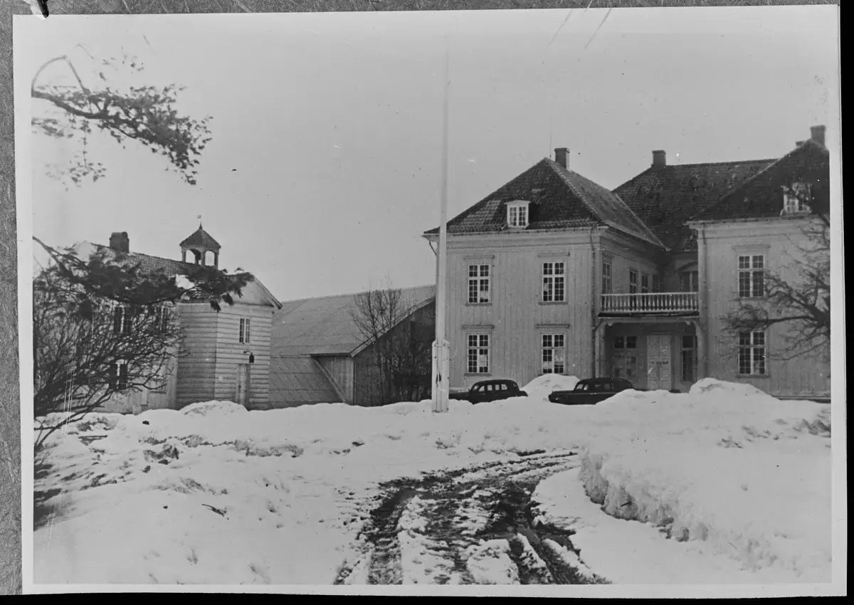 Eidsvollsbygningen omgitt av snø. Eidsvollsbygningen står sentralt i bildet, med en flaggstang uten flagg foran. Ujevn snø og hjulspor leder opp mot hovedinngangen, hvor noen biler er parkert.