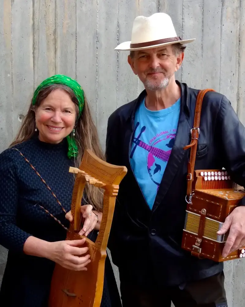 nærbilde av to musikere, en mann og en dame. Hun har langt hår og grønt pannebånd med harpelignende instrument. han har hatt og gammelt trekkspill over skuldra.