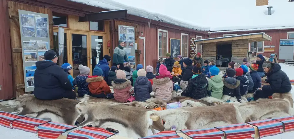 Mange barn sitter på benker med reinsdyrskinn i sirkel rundt foredragsholder. Utendørs i bakgård, snødekt landskap.