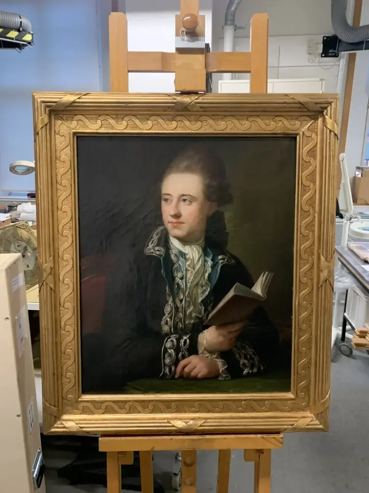 Portrettmaleri av Bernt Anker på en staffeli i et verksted. Anker er kledd i en mørk drakt med broderte detaljer. Han holder en bok og ser tenksomt til siden. Maleriet har en gullramme, og bakgrunnen viser et rom for kunsthåndtering.