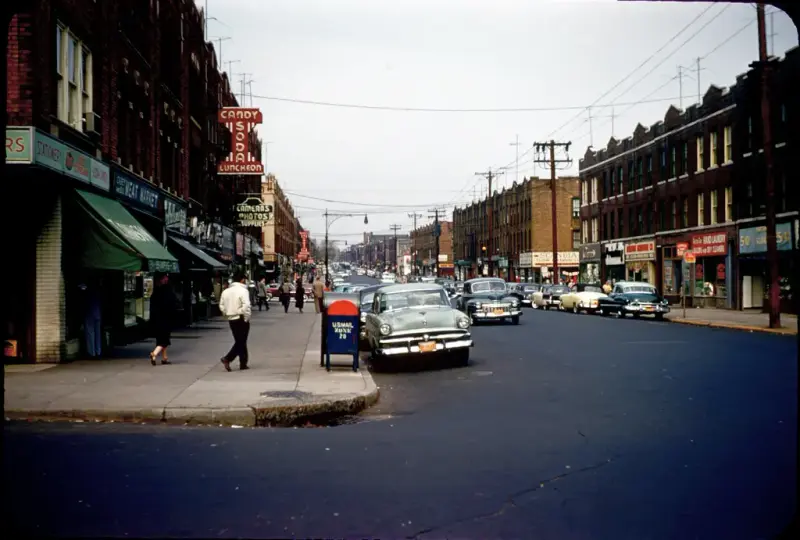 Gatebilde fra "Lapskaus Boulevard" i New York, med butikker, folk og klassiske biler.