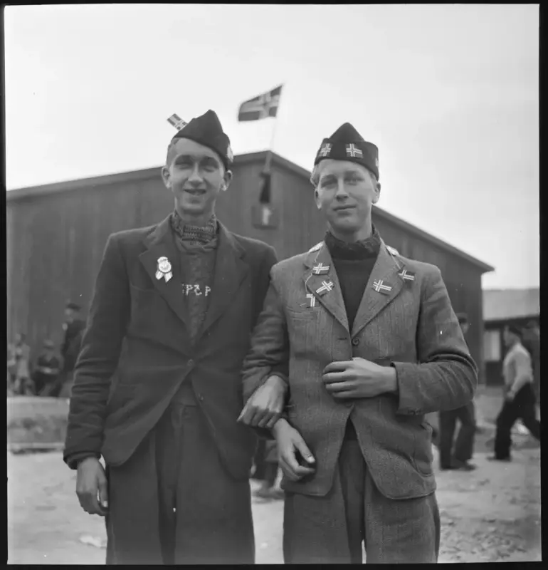 Svart/hvitt fotografi. To unge menn ser rett i kamera, de har på seg fangelue som er pyntet med norske flagg, og jakke som er pyntet med norske flagg. I bakgrunnen ser man en brakke hvor det svaier et norsk flagg.