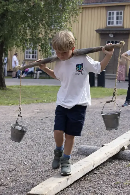 Boy balancing on a plank with milk pails over his shoulders during a children's activity at Norsk Folkemuseum.