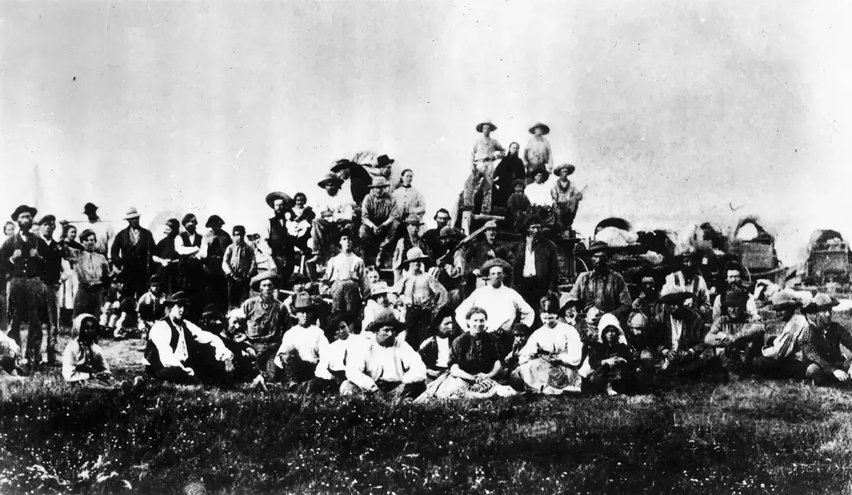 Historisk bilde av en stor gruppe mennesker. Overdekkede prærievogner bak til høyre.